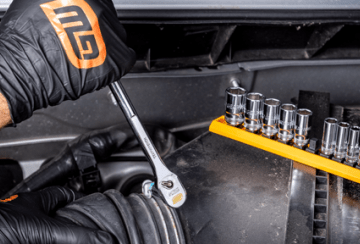 Man's hand wearing a black nitrile glove with GEARWRENCH logo using a 120XP ratchet to turn a fastener inside of an engine bay with a deep chrome socket set on a orange socket rail